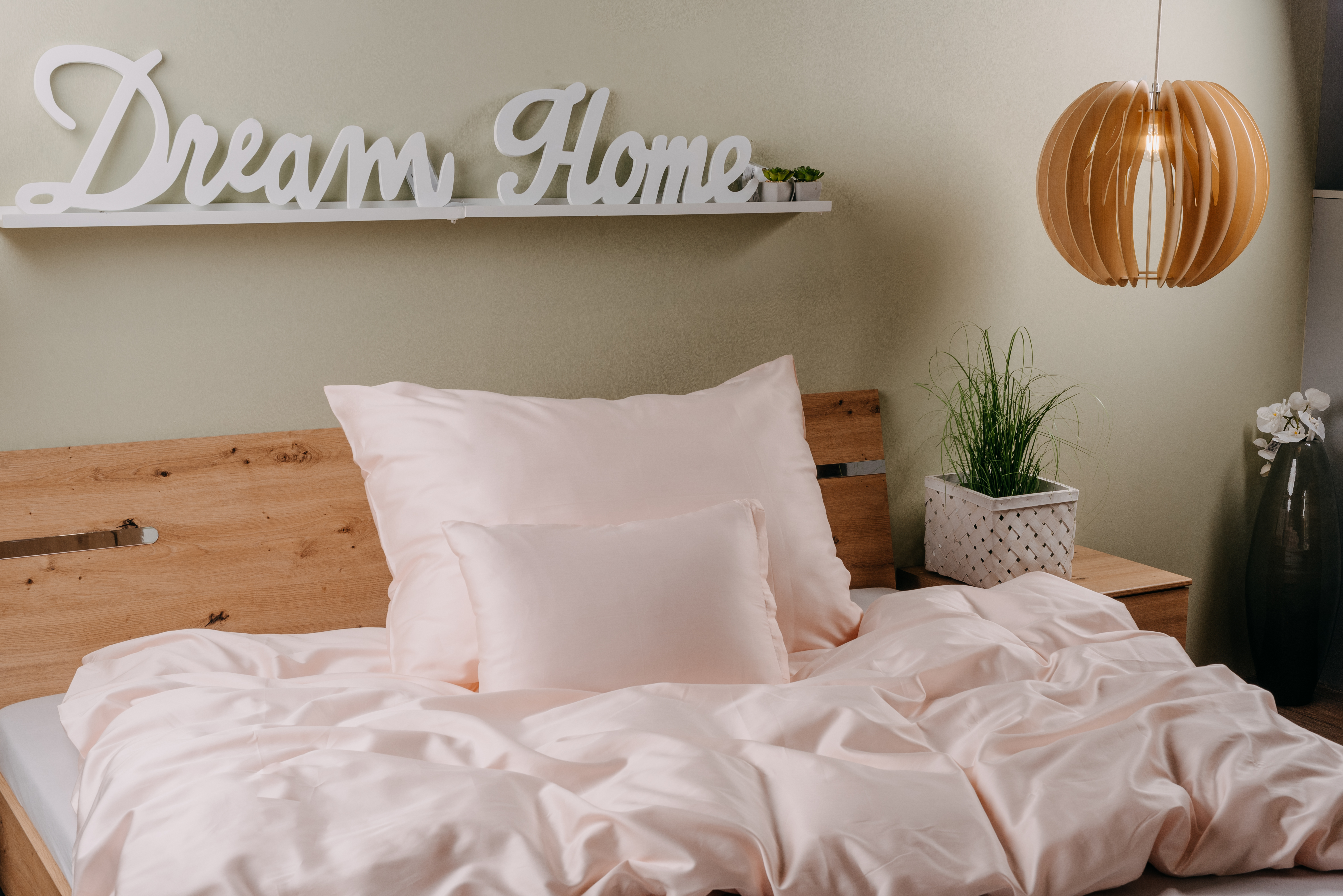 Hálószoba jelenet világos rózsaszín ágyneművel, fa fejtámlával és "Dream Home" felirattal.