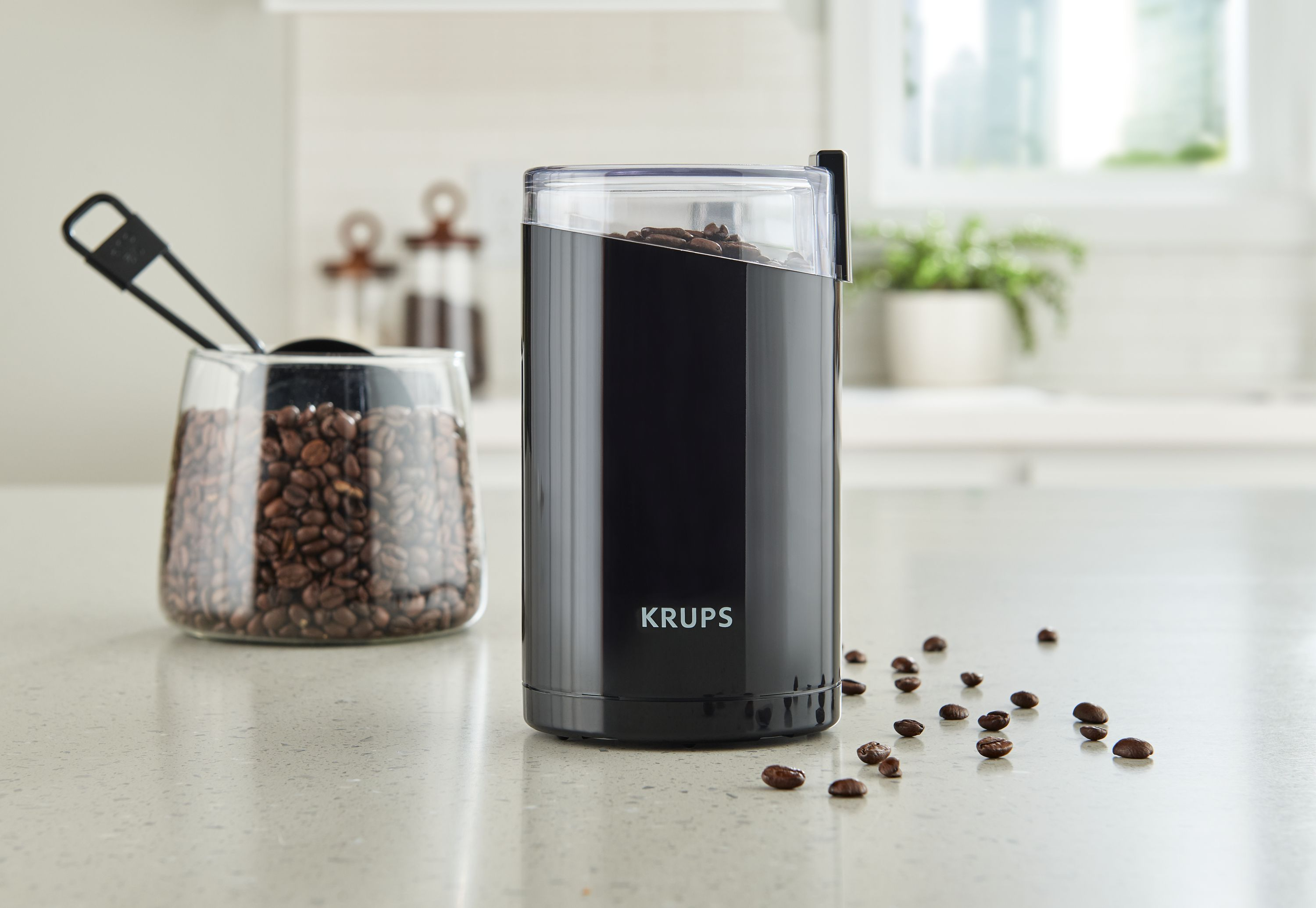 Schwarzer Krups-Kaffeemühle mit durchsichtigem Deckel, gefüllt mit Bohnen. Glasbehälter mit Bohnen links.