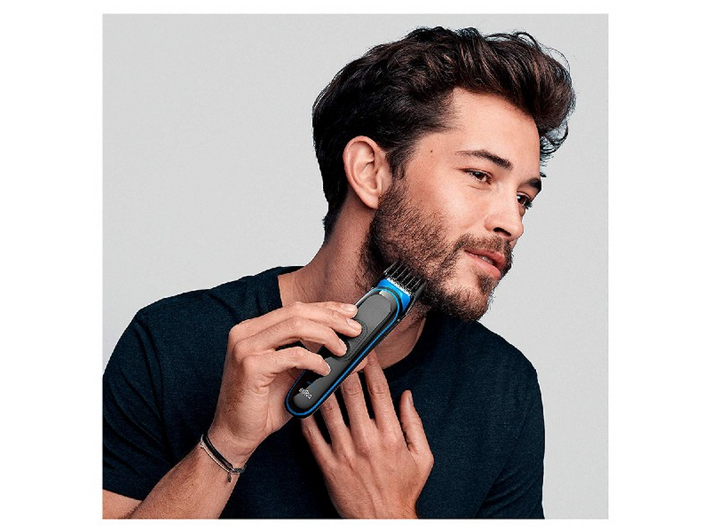 Un hombre recortando su barba con una recortadora de barba negra y azul. Lleva una camisa oscura.