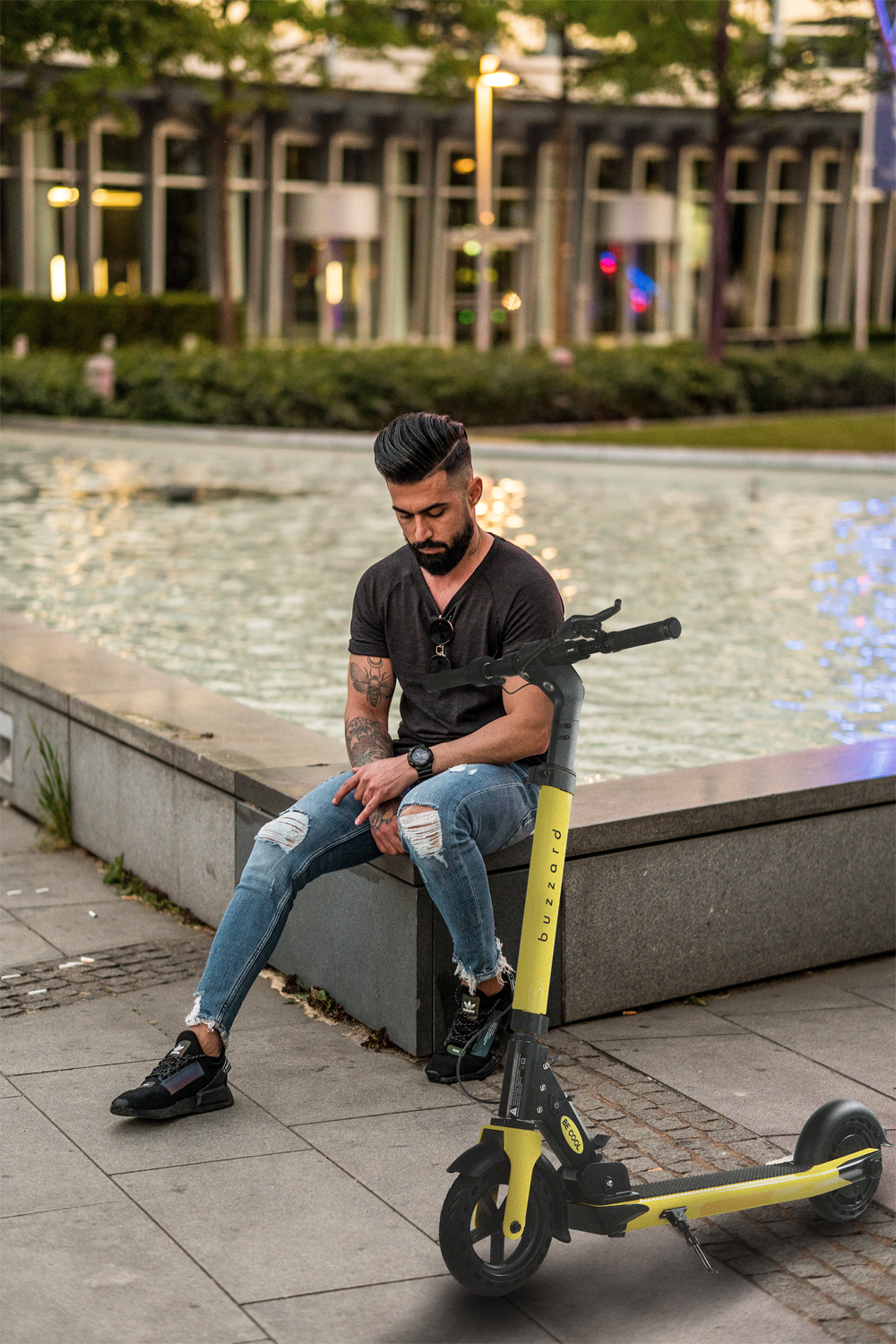 Mann sitzt neben einem gelben Roller neben einem Brunnen. Er trägt Jeans und ein schwarzes Hemd.