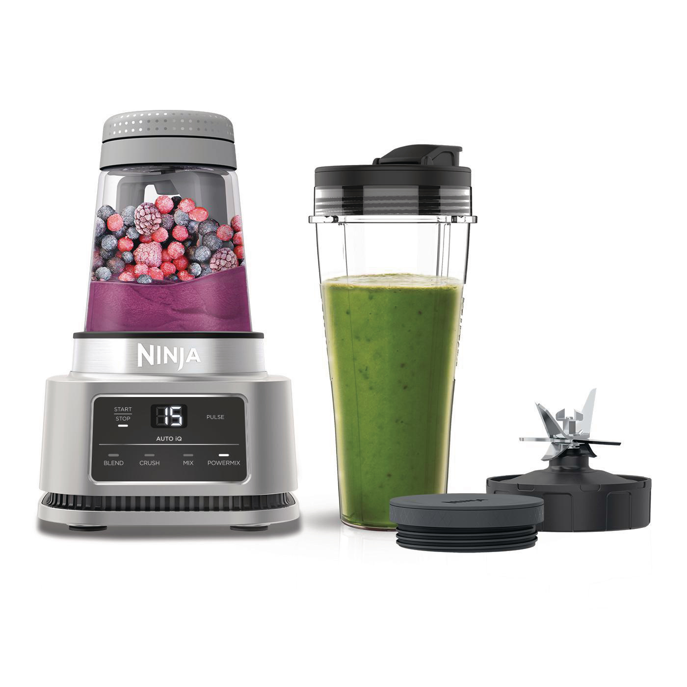 Ninja-Mixer mit Smoothie-Zutaten. Ein volles Glas und ein Becher mit Deckel sind vorhanden. Schwarze Klinge.