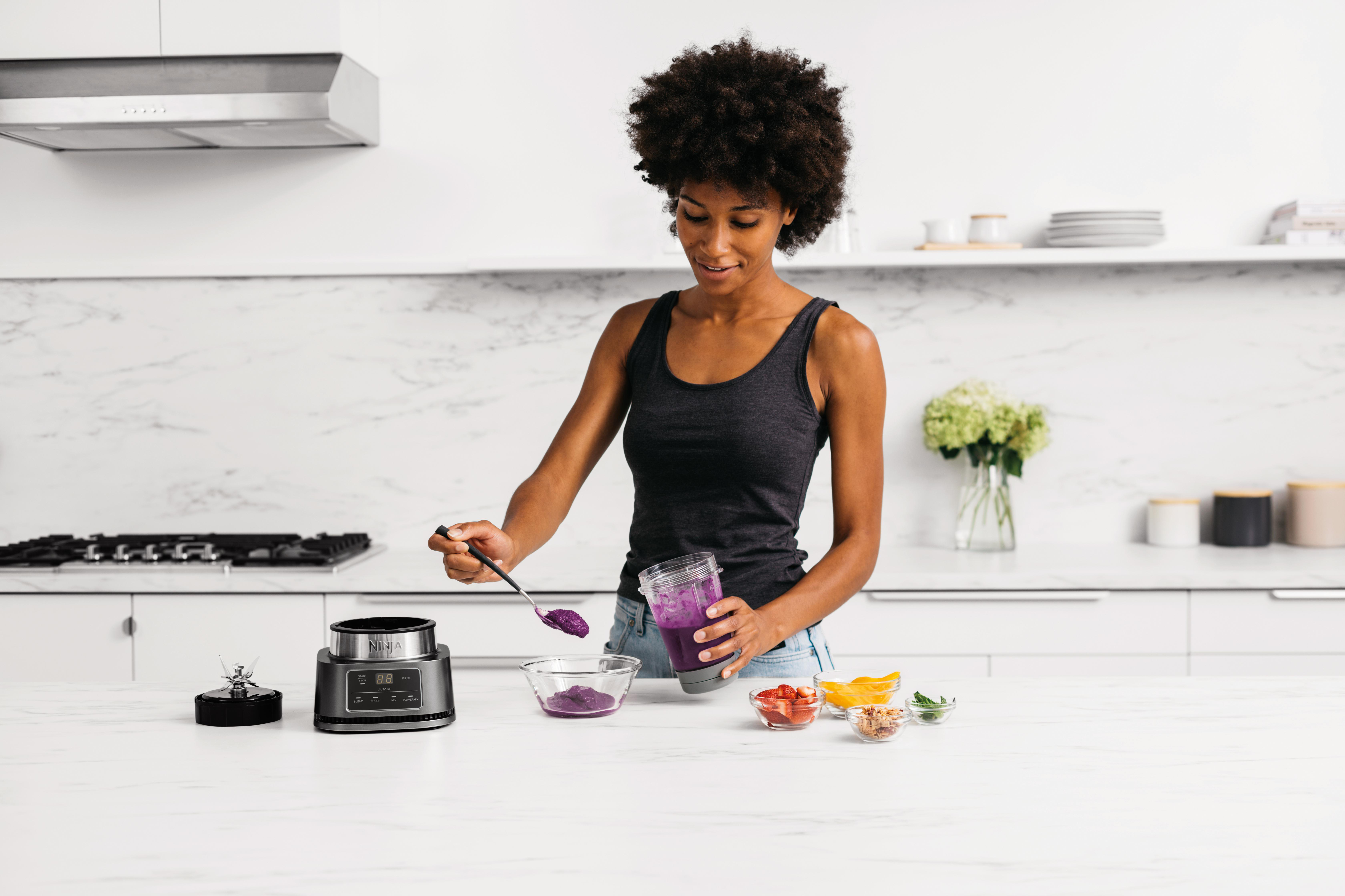 Frau, die einen Smoothie zubereitet, ihn in eine Schüssel gießt, mit Mixer und Zutaten auf der Theke.