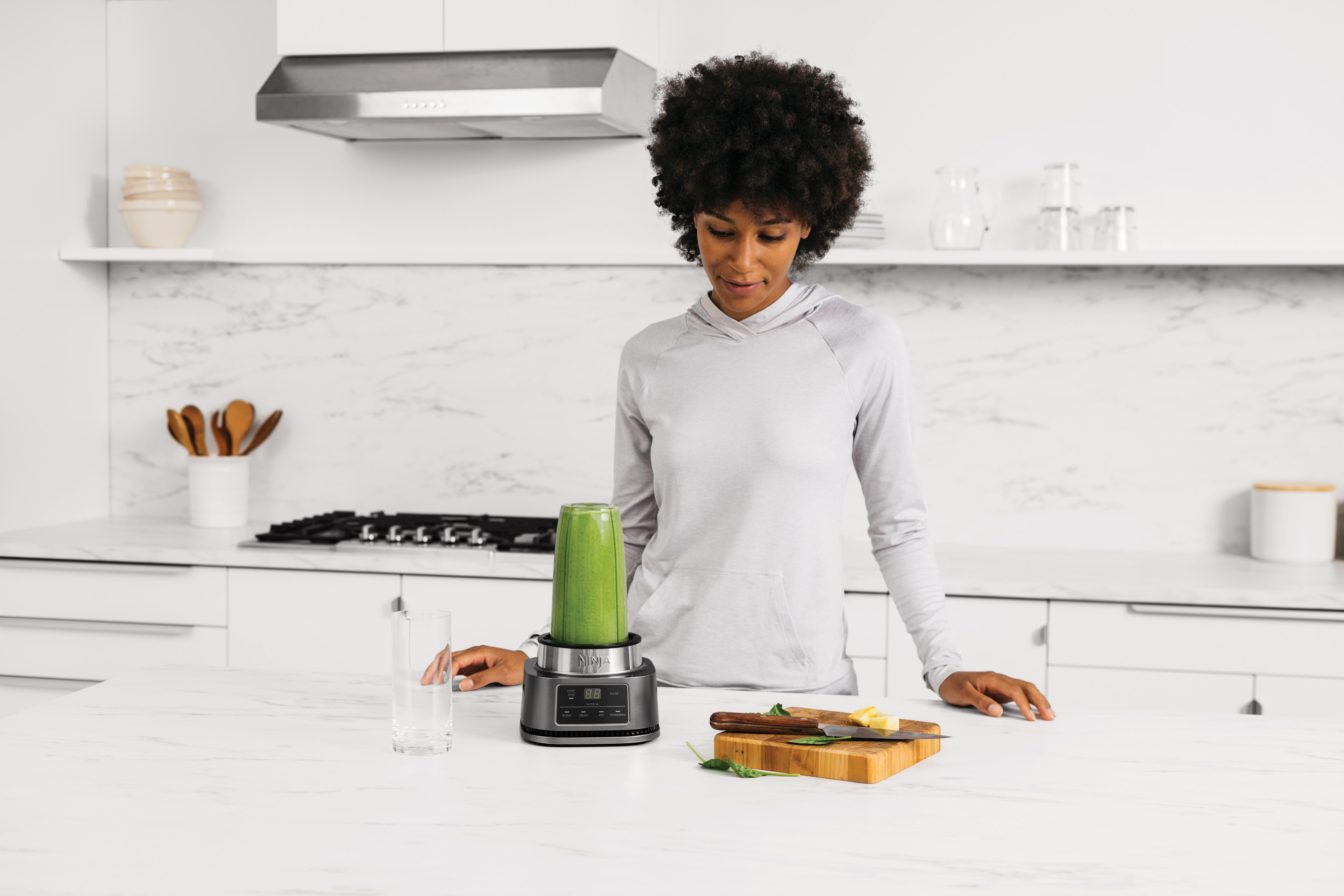 Eine Frau macht einen Smoothie mit einem Mixer. Zutaten liegen auf einem Schneidebrett daneben.