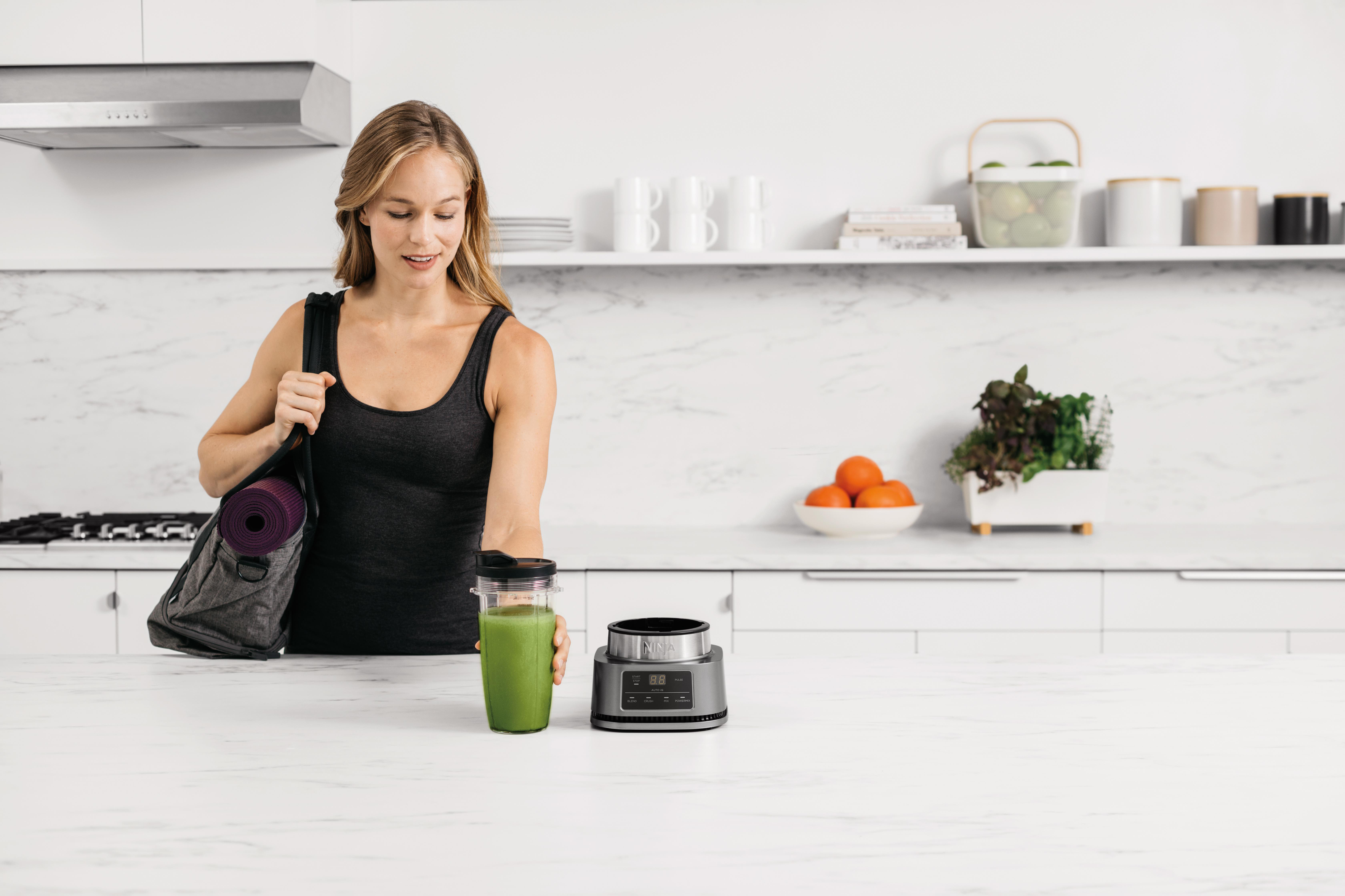 Eine Frau mit Sporttasche und Smoothie steht in einer weißen Küche. Es gibt Früchte und Pflanzen.