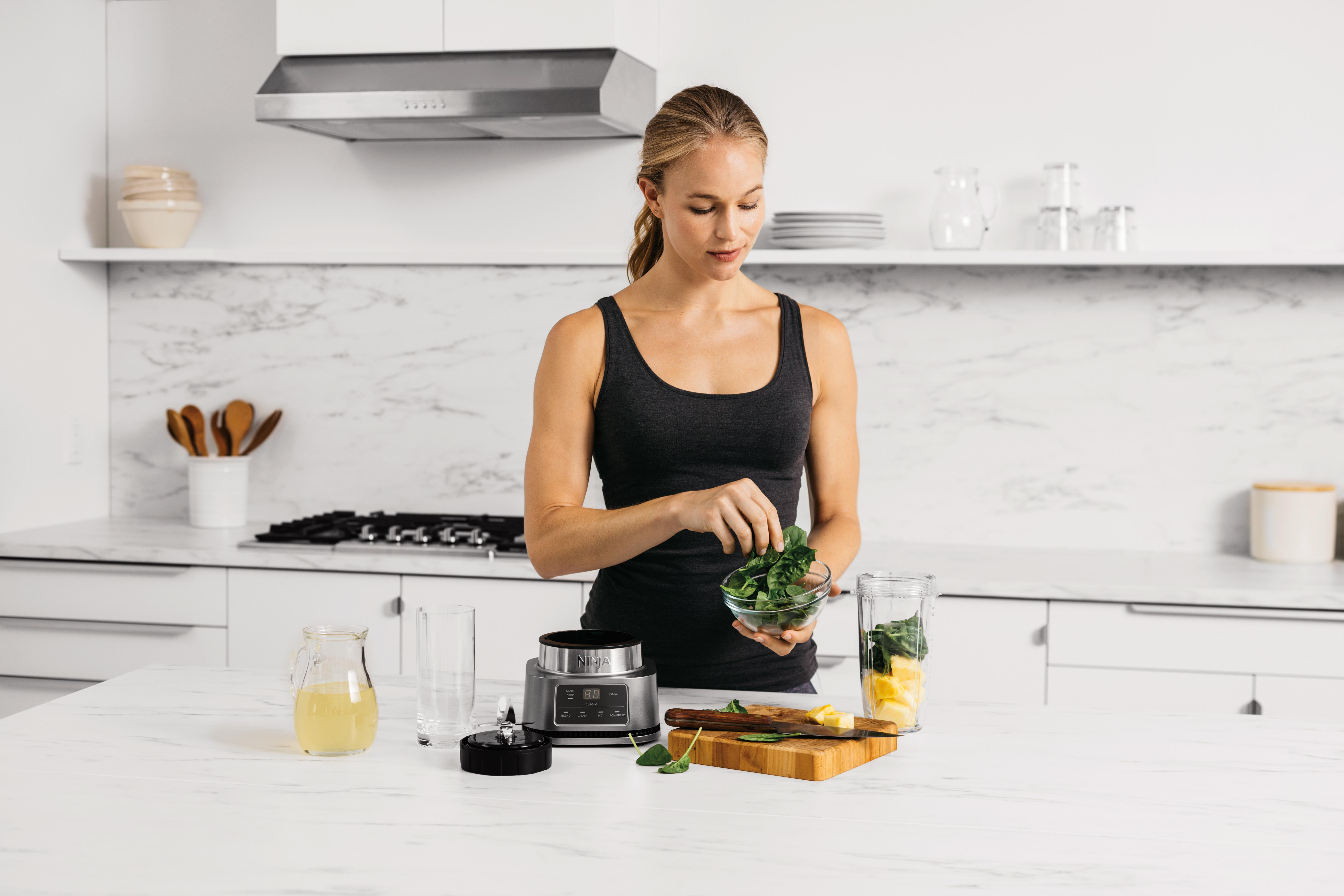 Frau, die einen Smoothie zubereitet, fügt Grünzeug in den Mixer. Küchenumgebung mit Zutaten.