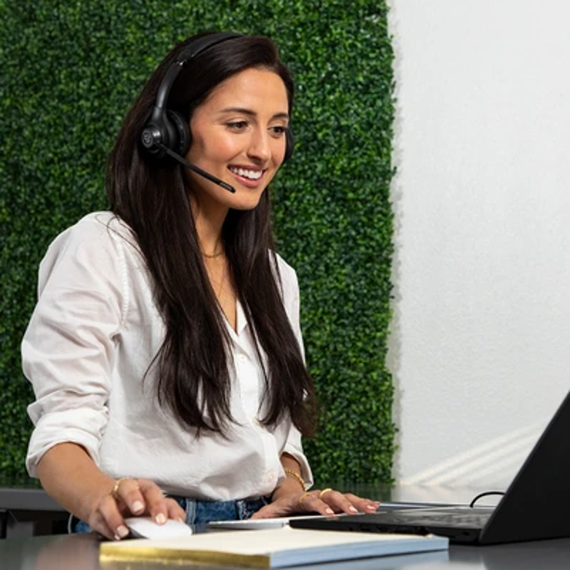Una donna con le cuffie sorride mentre guarda un laptop. Sullo sfondo c'è un muro verde.