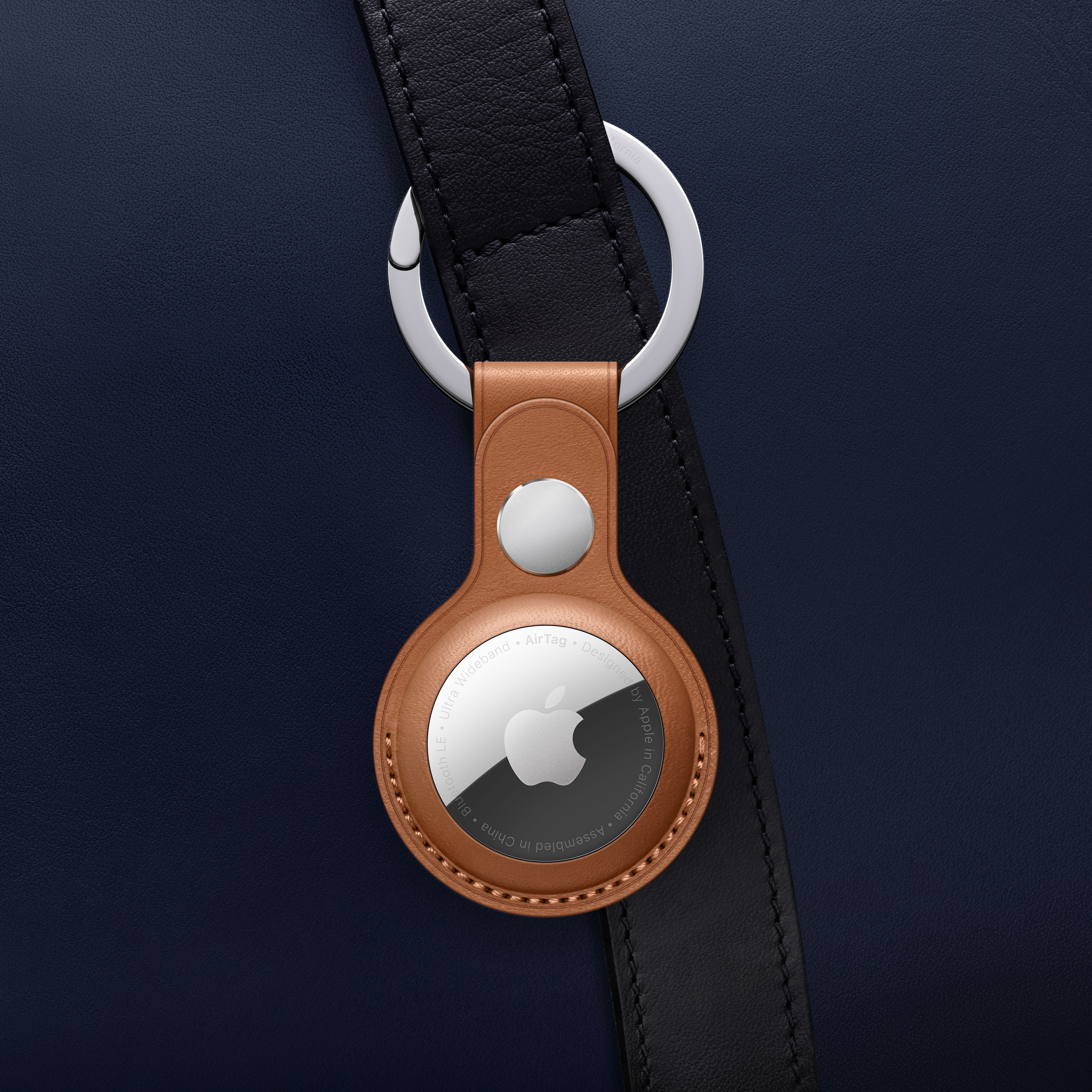 Apple AirTag an einem braunen Lederhalter. Es ist an einem schwarzen Riemen auf blauem Grund befestigt.