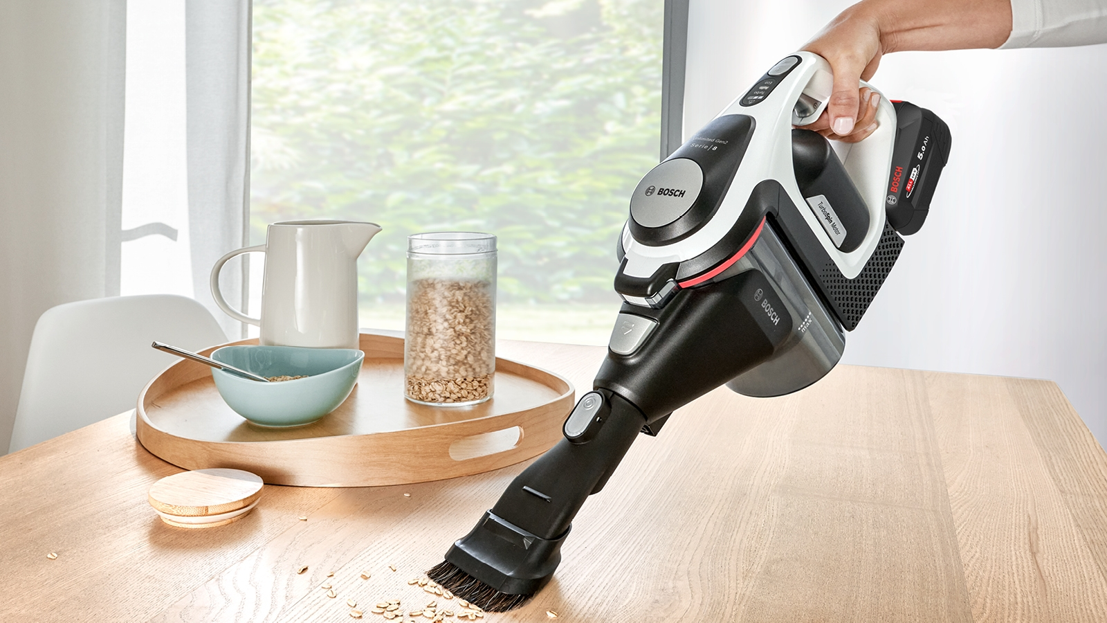 Eine Frau benutzt einen kabellosen Staubsauger, um einen Tisch zu reinigen. Ein Holztablett ist sichtbar.