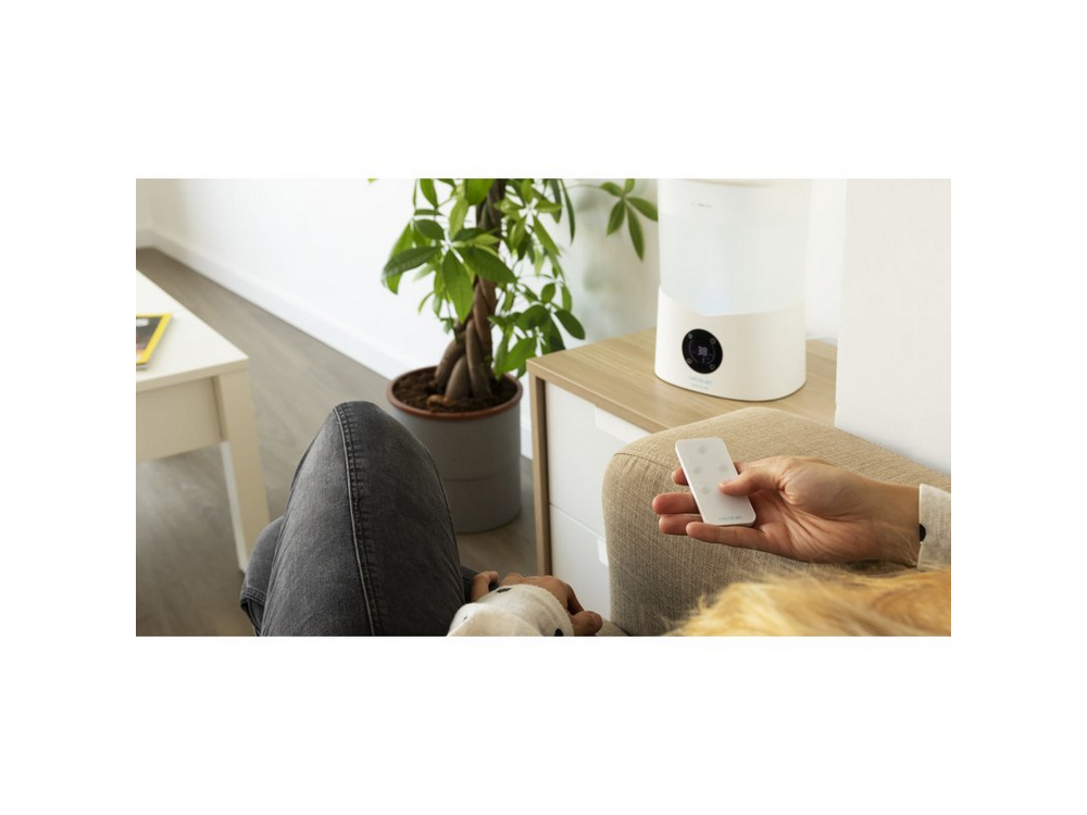 Una persona en un sofá sostiene un mando a distancia blanco. Un humidificador y una planta están en el fondo.