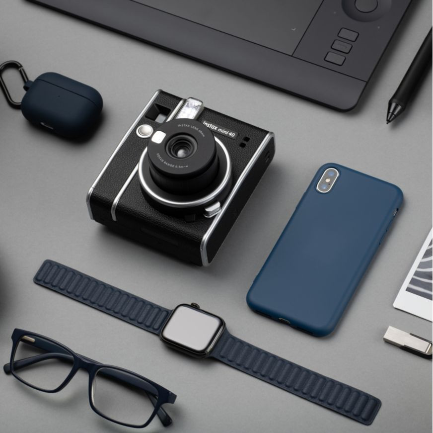 Een zwarte camera, telefoon, horloge, bril en accessoires op een grijs oppervlak.