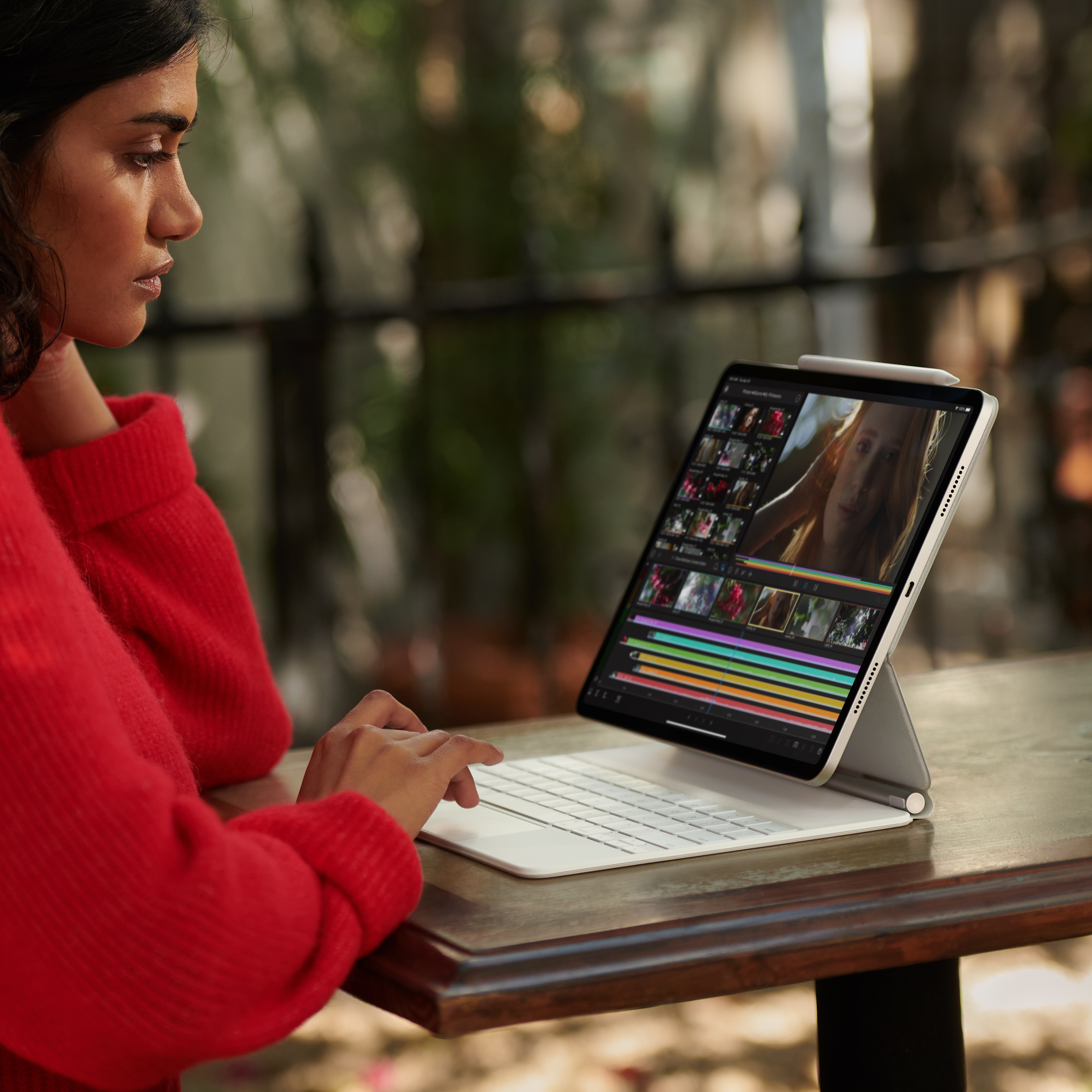 Mujer usando una tableta con teclado en una mesa exterior. La pantalla muestra interfaz de edición de video.
