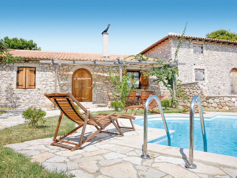 Casa in pietra con piscina. Sedie e lettini in legno. Cielo blu e acqua limpida.