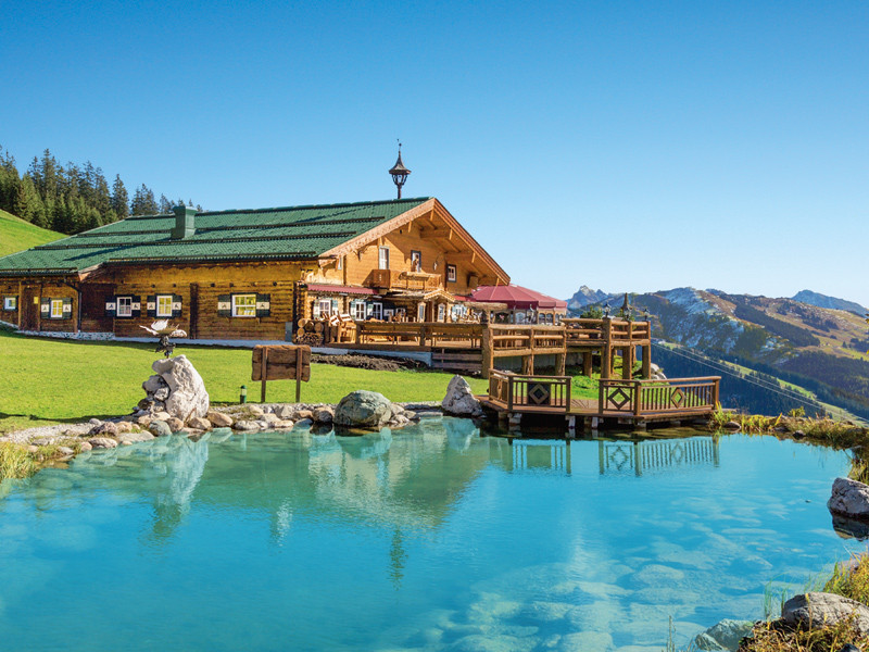 Una baita in legno vicino a un lago tra le montagne in una giornata di sole. Tetto verde e acqua blu.