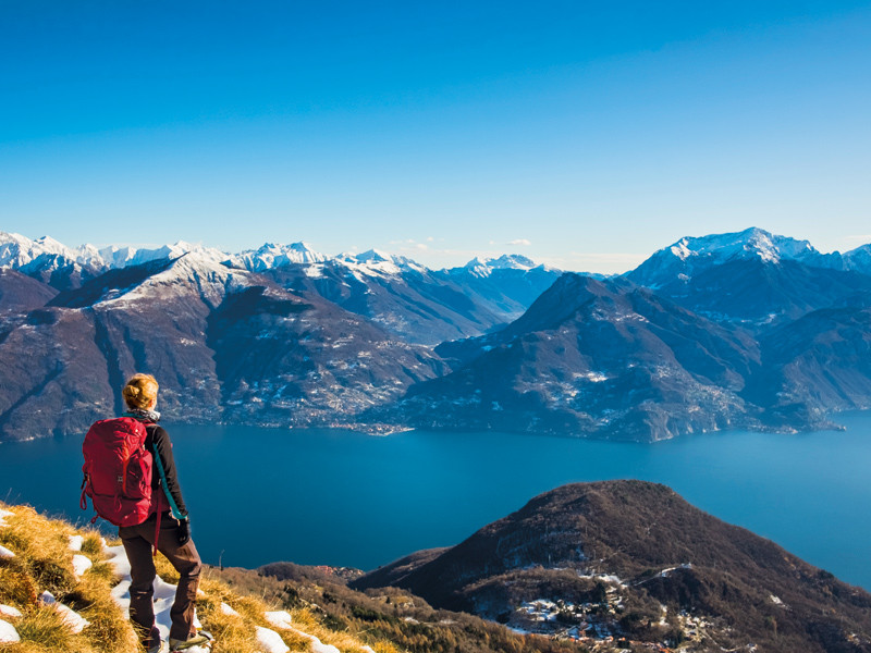 Una donna con uno zaino rosso guarda un lago circondato da montagne innevate sotto un cielo blu.