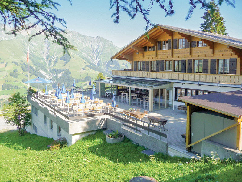 Edificio in legno con terrazza, tavoli e sedie, con montagne sullo sfondo.