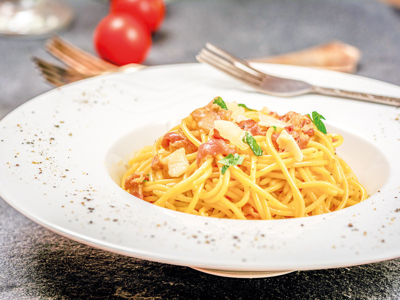Spaghetti alla carbonara in una ciotola bianca, con pomodori e una forchetta sullo sfondo, con un po' di pepe nero.