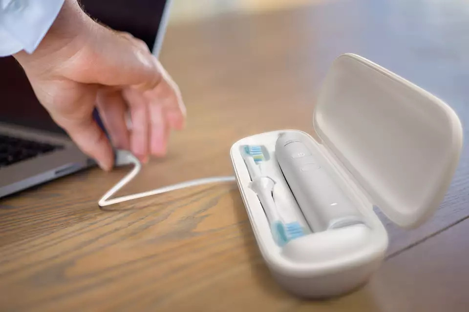 A white electric toothbrush and case are connected to a laptop via a charging cable on a wooden surface.