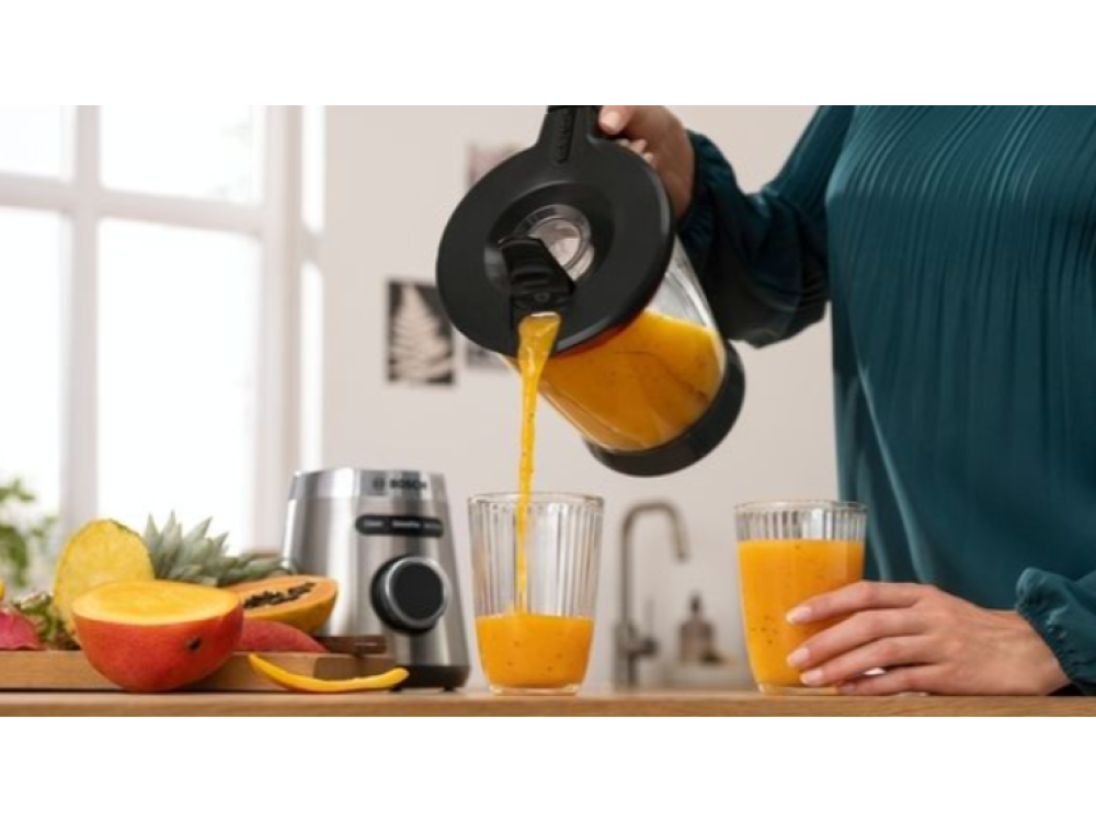 Una persona vertiendo jugo de naranja de una jarra en dos vasos, con frutas y una licuadora en el fondo.