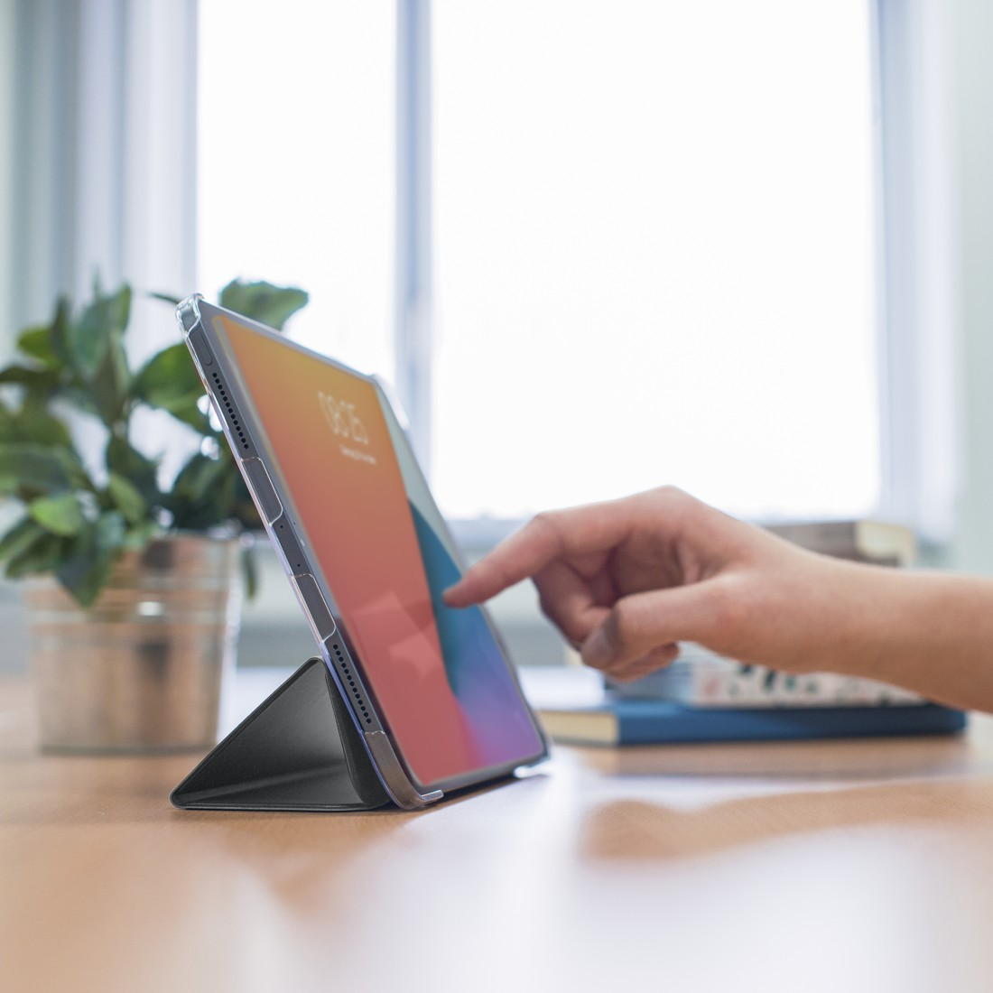 Tablet auf Ständer, Hand berührt Bildschirm, Bücher und Pflanze im Hintergrund, in der Nähe eines Fensters.