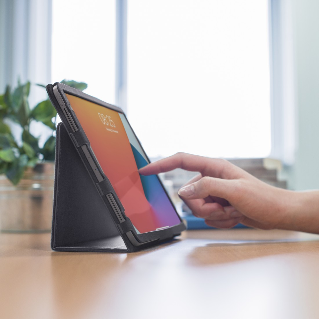 Ein iPad in einem Gehäuse liegt auf einem Schreibtisch. Eine Hand berührt den Bildschirm. Der Bildschirm zeigt Orange.