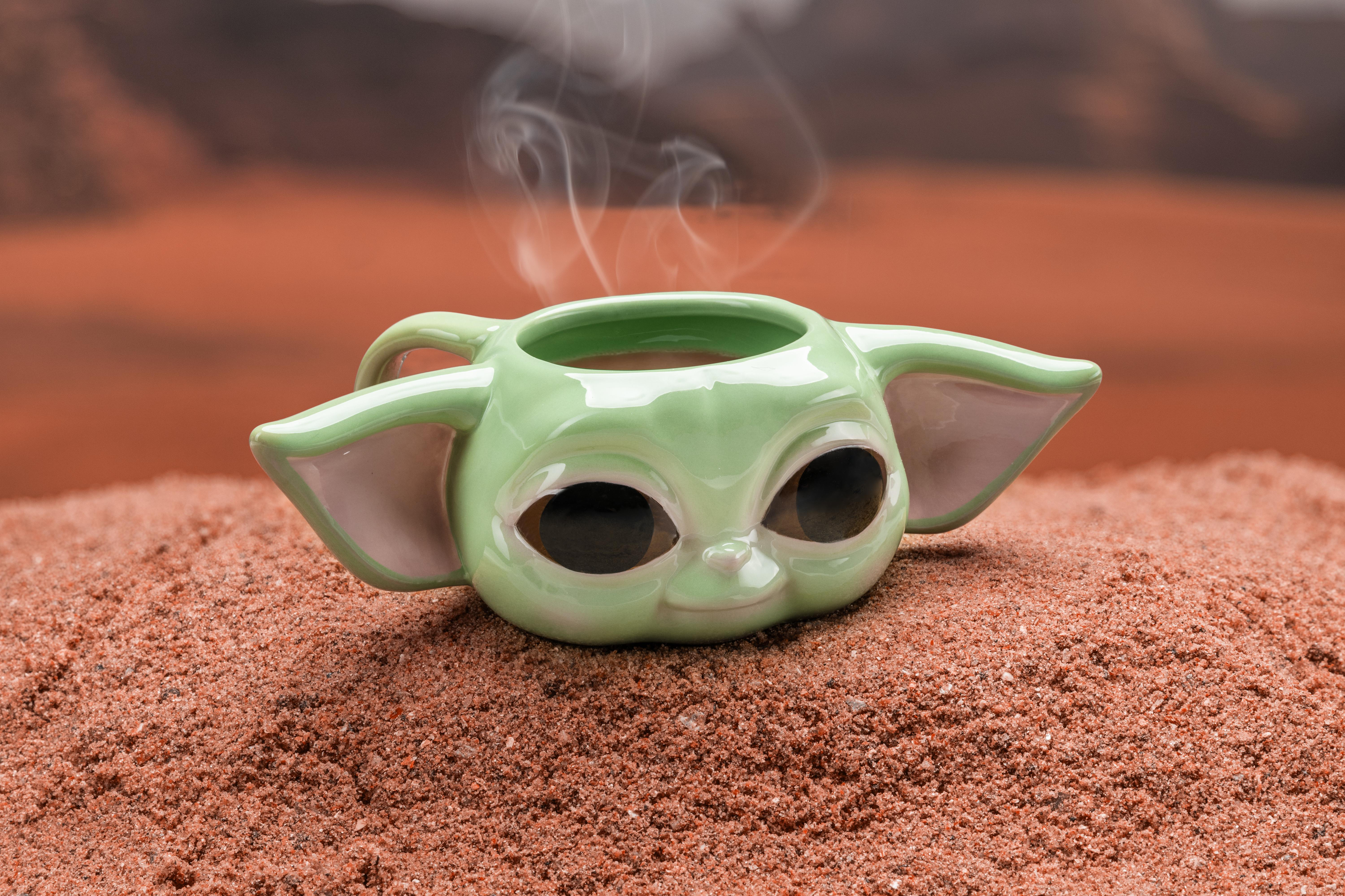 Ein grüner Grogu-Becher mit aufsteigendem Dampf, auf sandiger Oberfläche mit rötlichem Hintergrund.