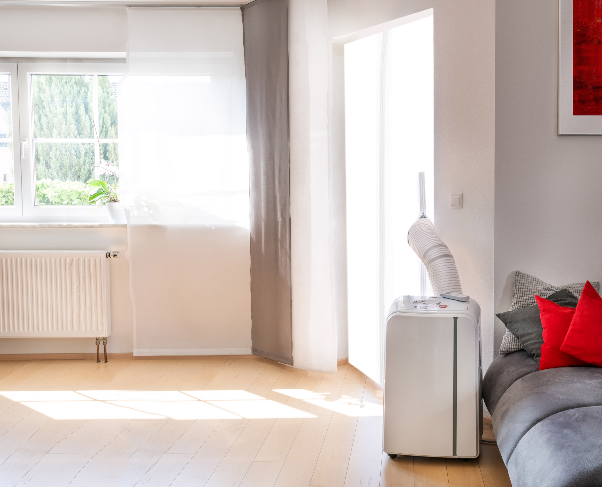 Un salon avec un climatiseur mobile, une fenêtre, un canapé et un sol en bois. Il y a de la lumière du soleil.