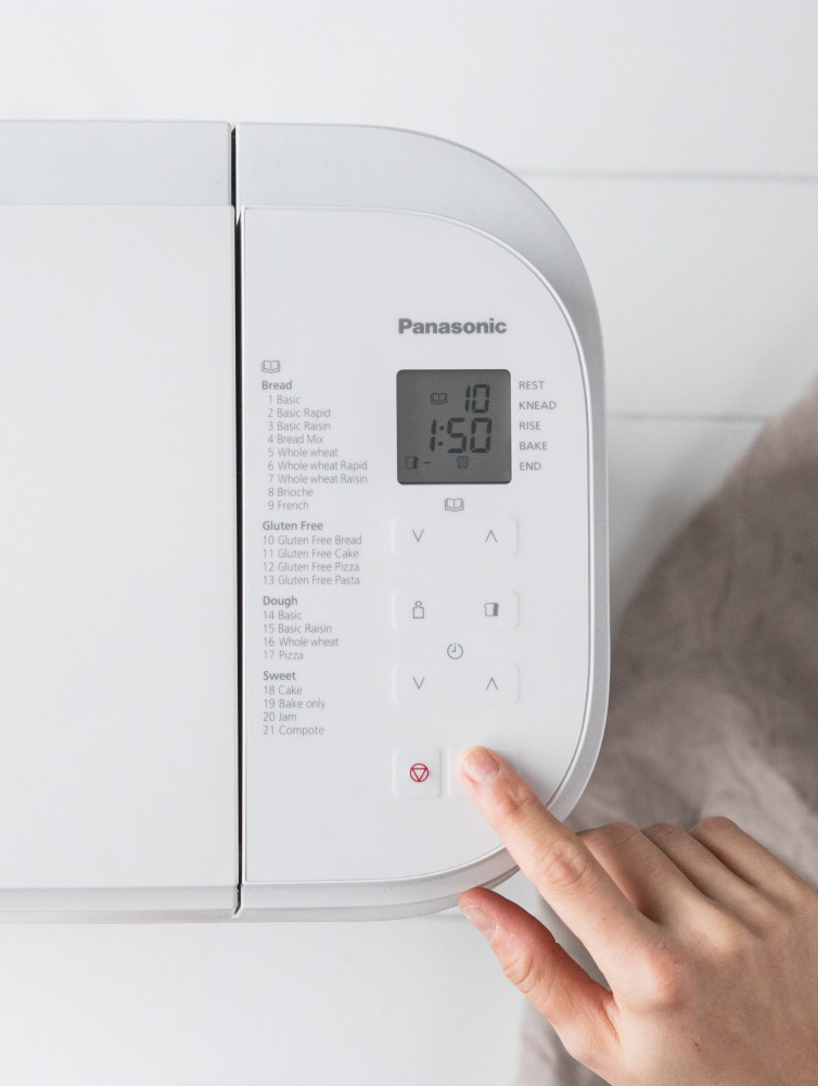 Fehér Panasonic kenyérsütő, kézzel nyomva egy gombot.