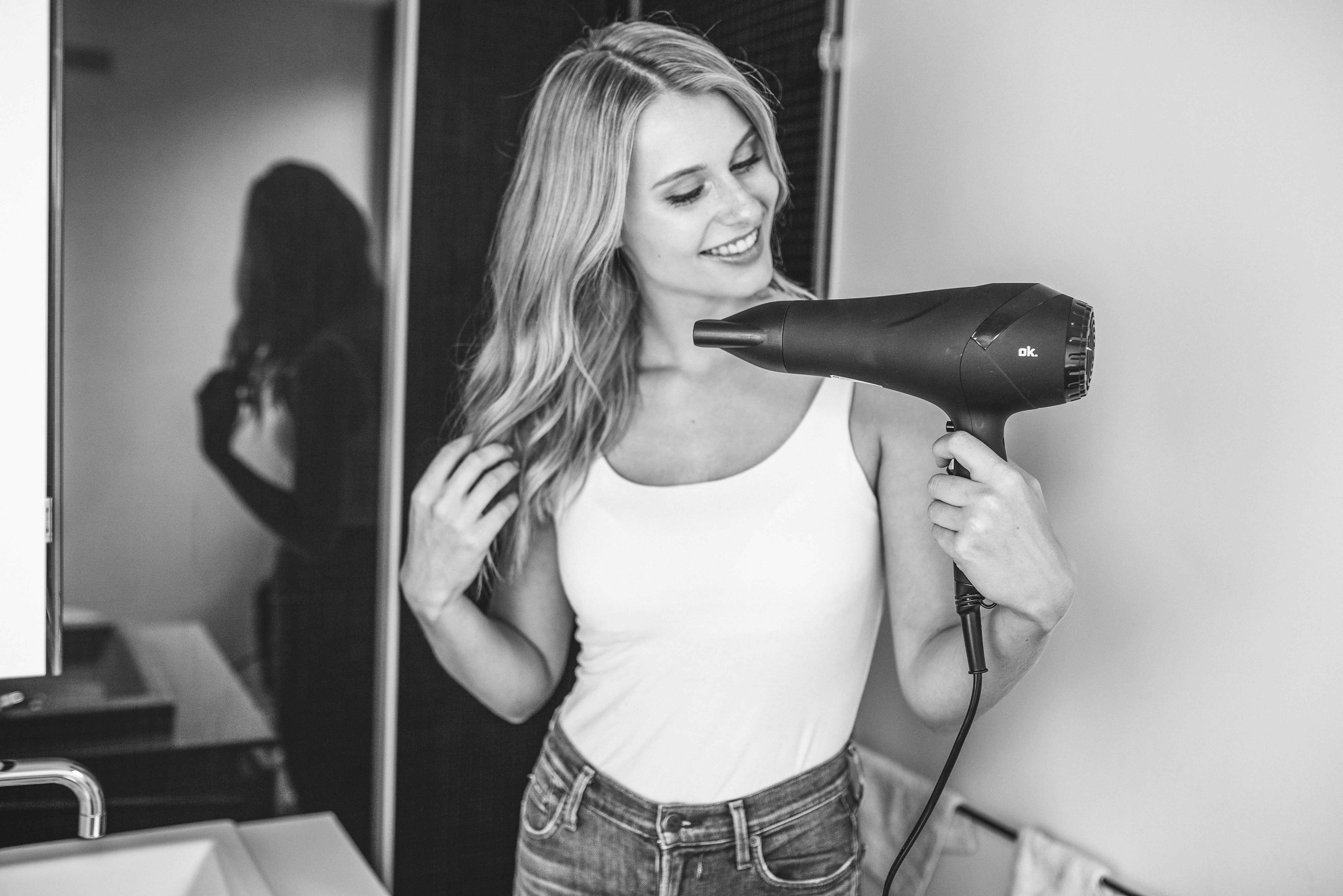 Blonde Frau lächelt, hält einen schwarzen Haartrockner. Sie trägt ein weißes Tanktop und Jeans, Badezimmer.