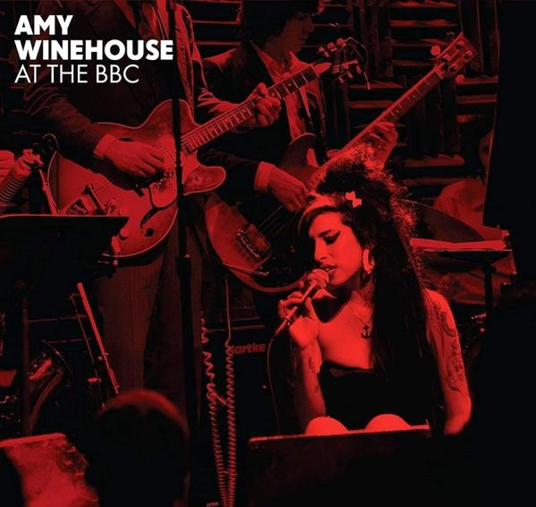 Amy Winehouse op het podium aan het zingen. Band speelt op de achtergrond. Rode en zwarte verlichting.