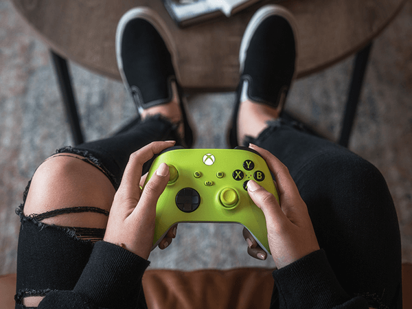 Yeşil bir Xbox denetleyicisi tutan kişi. Bacaklar ve siyah ayakkabılar görünür. Sandalyede oturuyor, yukarıdan görünüm.