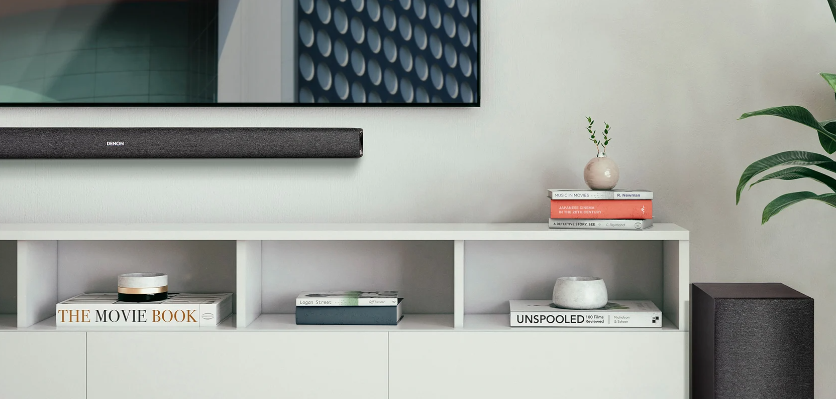 Nappali TV-vel, soundbarral, könyvekkel és növénnyel, fehér fal előtt.