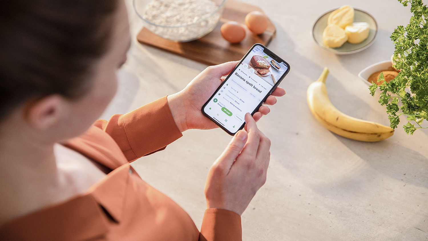 Een persoon die een smartphone vasthoudt met een recept op het scherm, omringd door ingrediënten op een aanrecht.