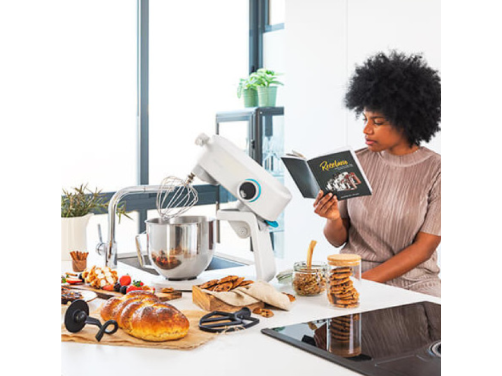 Mujer leyendo un libro en la cocina con una batidora y comida en la encimera.