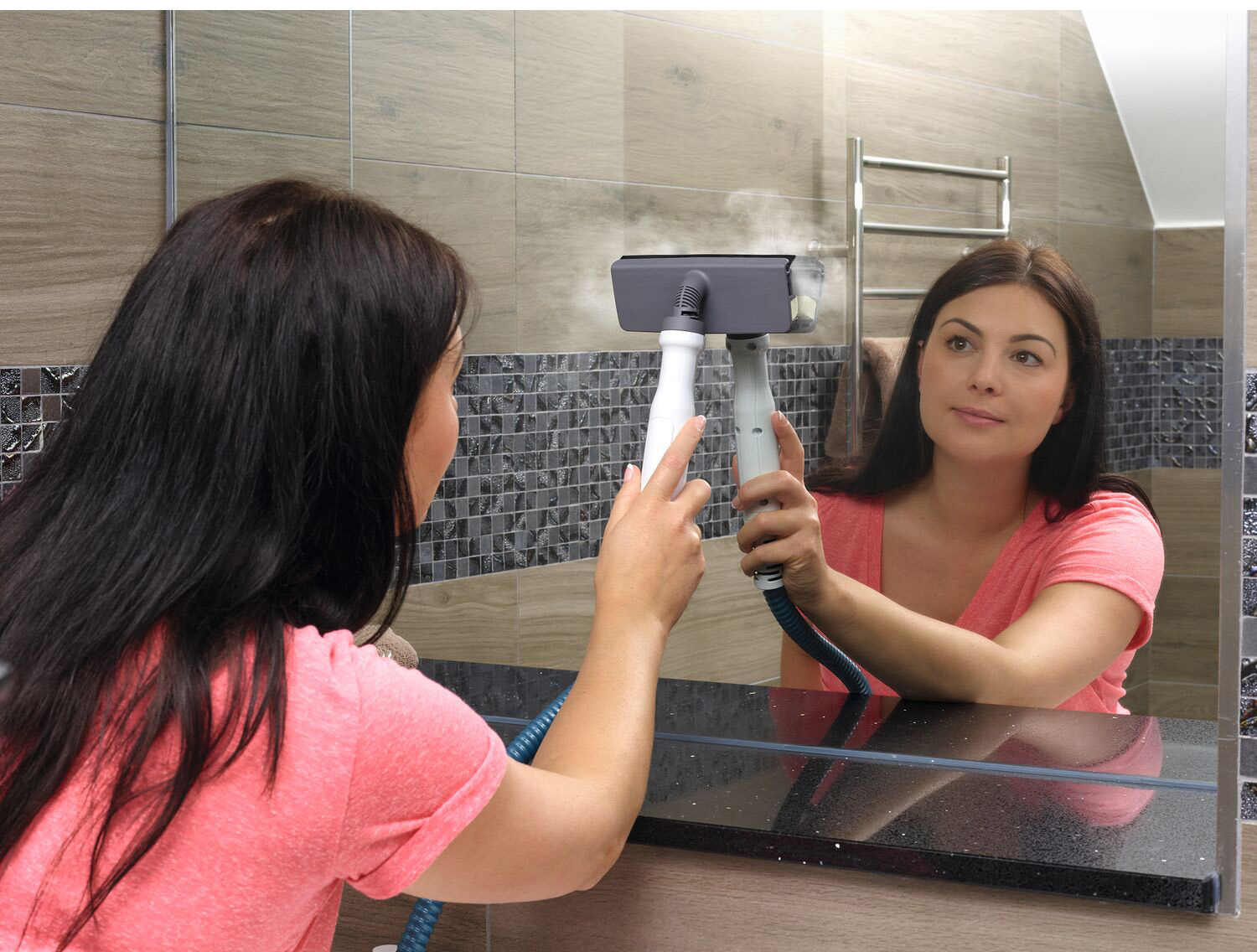 Woman cleaning a mirror with a steamer. Mirror is reflecting the woman. Beige and gray tiles.