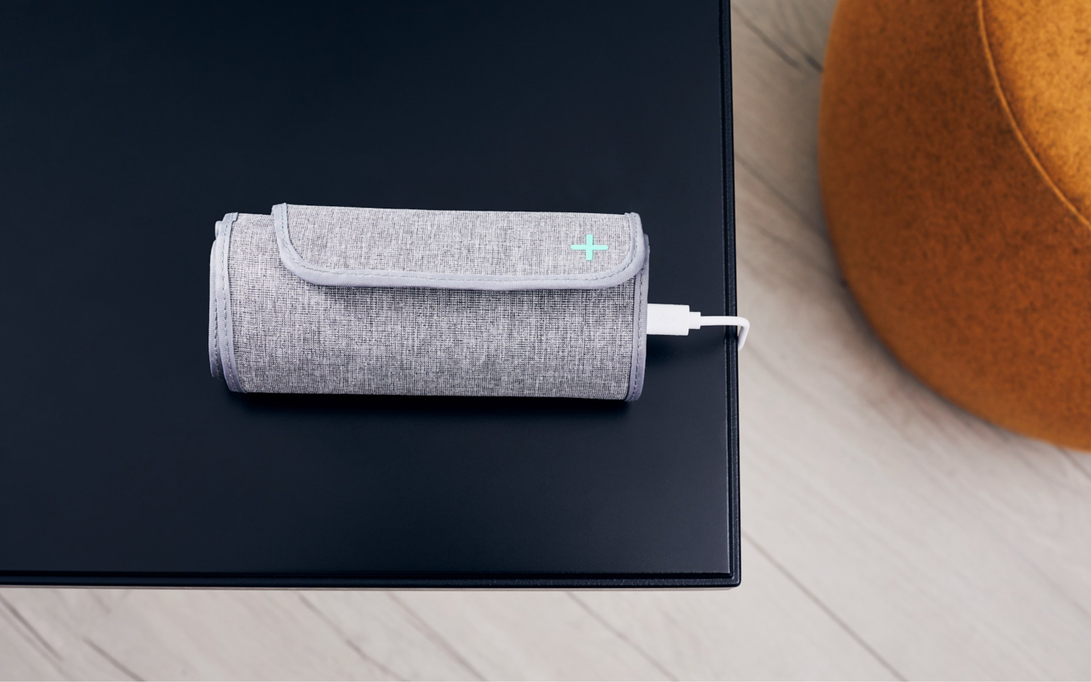 Un appareil médical gris avec un signe plus, relié à un câble blanc, est posé sur une table noire. Un pouf orange en arrière-plan.