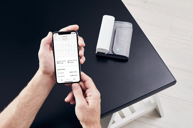 Une personne tient un smartphone affichant des données de pression artérielle. Un appareil blanc et un brassard gris sont sur une table noire.