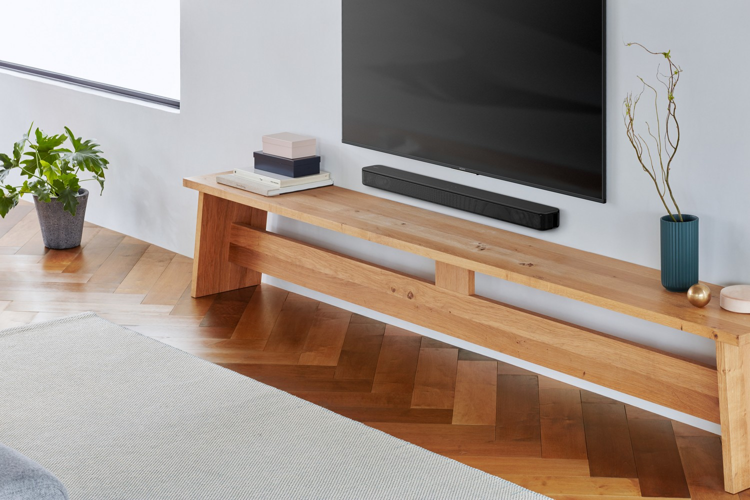 Escena de salón con banco de madera, TV, barra de sonido, plantas y alfombra clara.