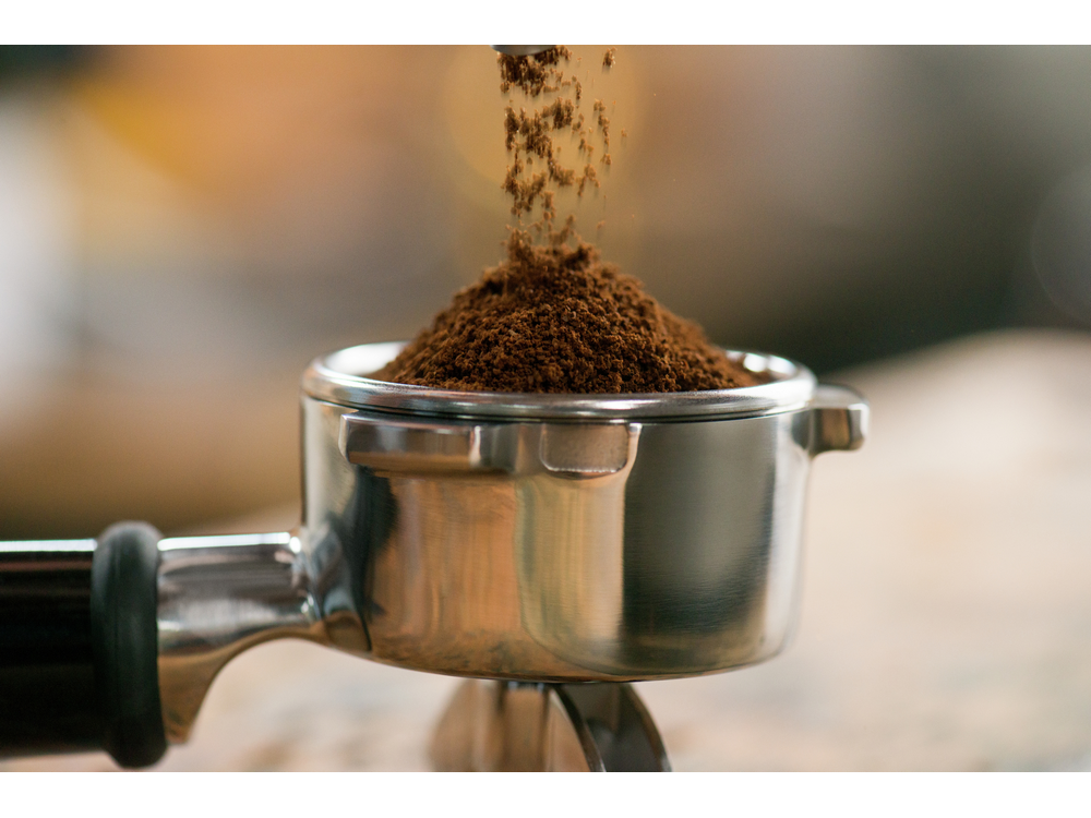 Un portafiltro plateado con granos de café vertiéndose en él, sobre un fondo borroso.