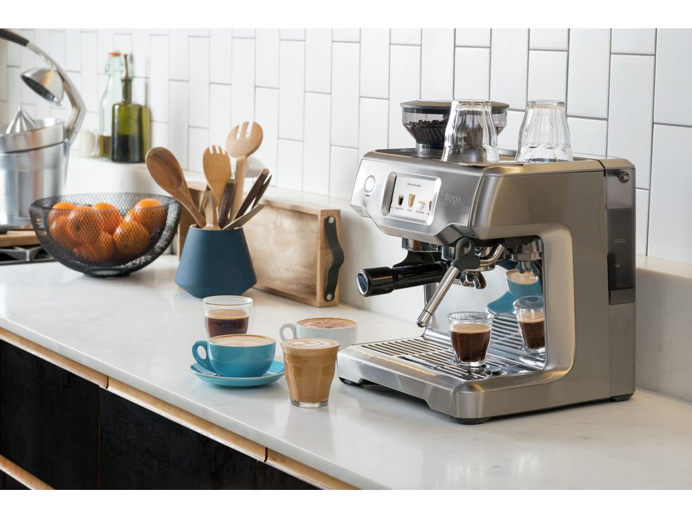 Una máquina de espresso plateada con varias tazas y un cuenco de fruta en una encimera de cocina.