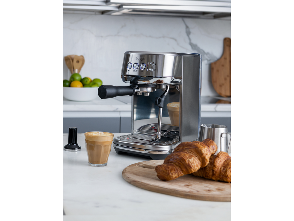 Una máquina de espresso de acero inoxidable con un vaso de latte y cruasanes en una tabla de madera sobre una encimera blanca.