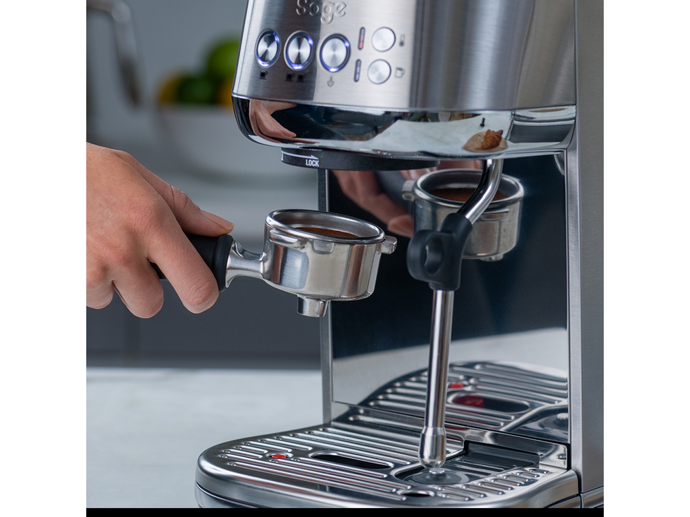 Una mano sostiene un portafiltro con café, máquina de espresso de acero inoxidable al fondo, ambiente de cocina.