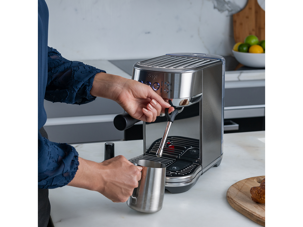 Persona usando una máquina de espresso plateada, sosteniendo una taza. Ambiente de cocina.