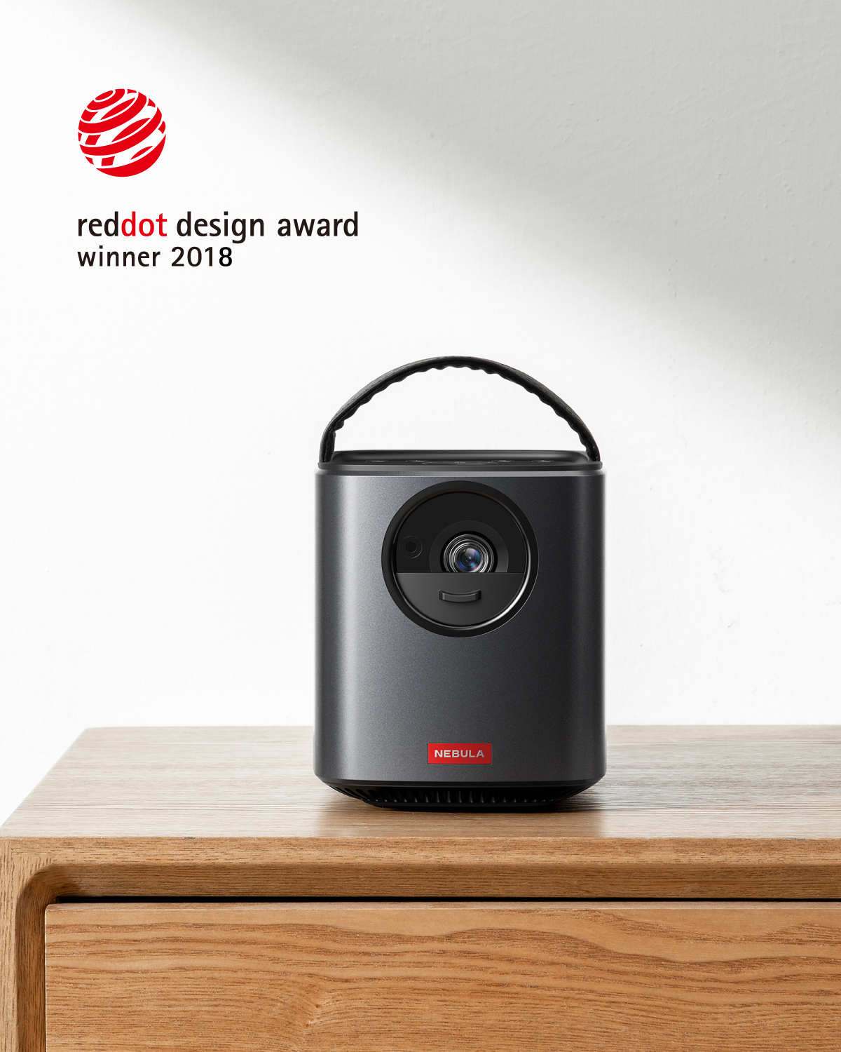 Ahşap bir masa üzerinde küçük, koyu gri bir projektör. Reddot ödül logosu g örünür.