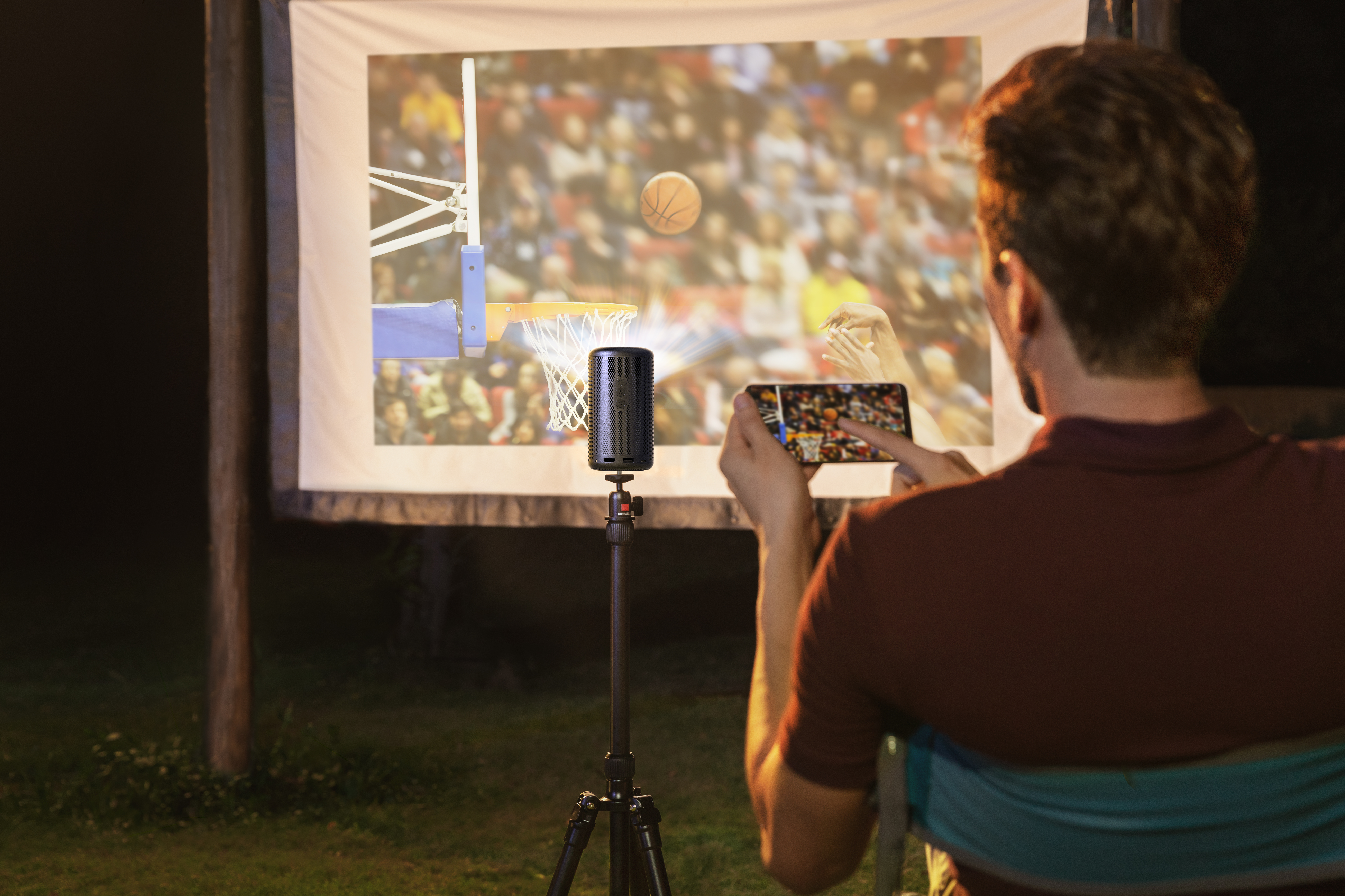 Bir kişi telefon tutuyor, bir ekranda yansıtılan bir basketbol maçını filme alıyor. Projektör bir tripoda yerleştirilmiş.