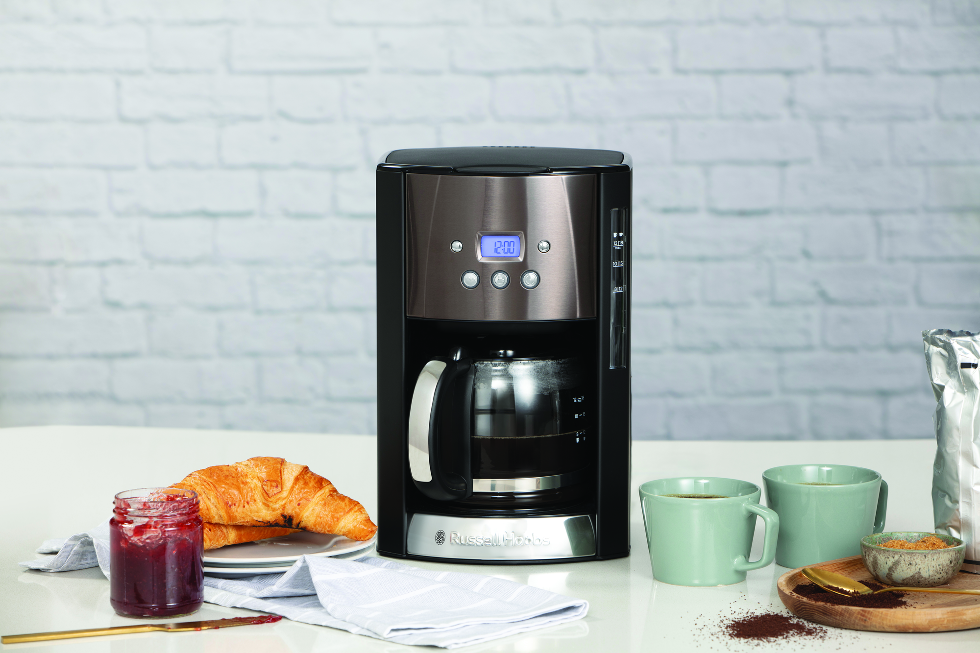 Coffee maker with croissants and jam on a table, mugs, and ingredients, white brick wall.