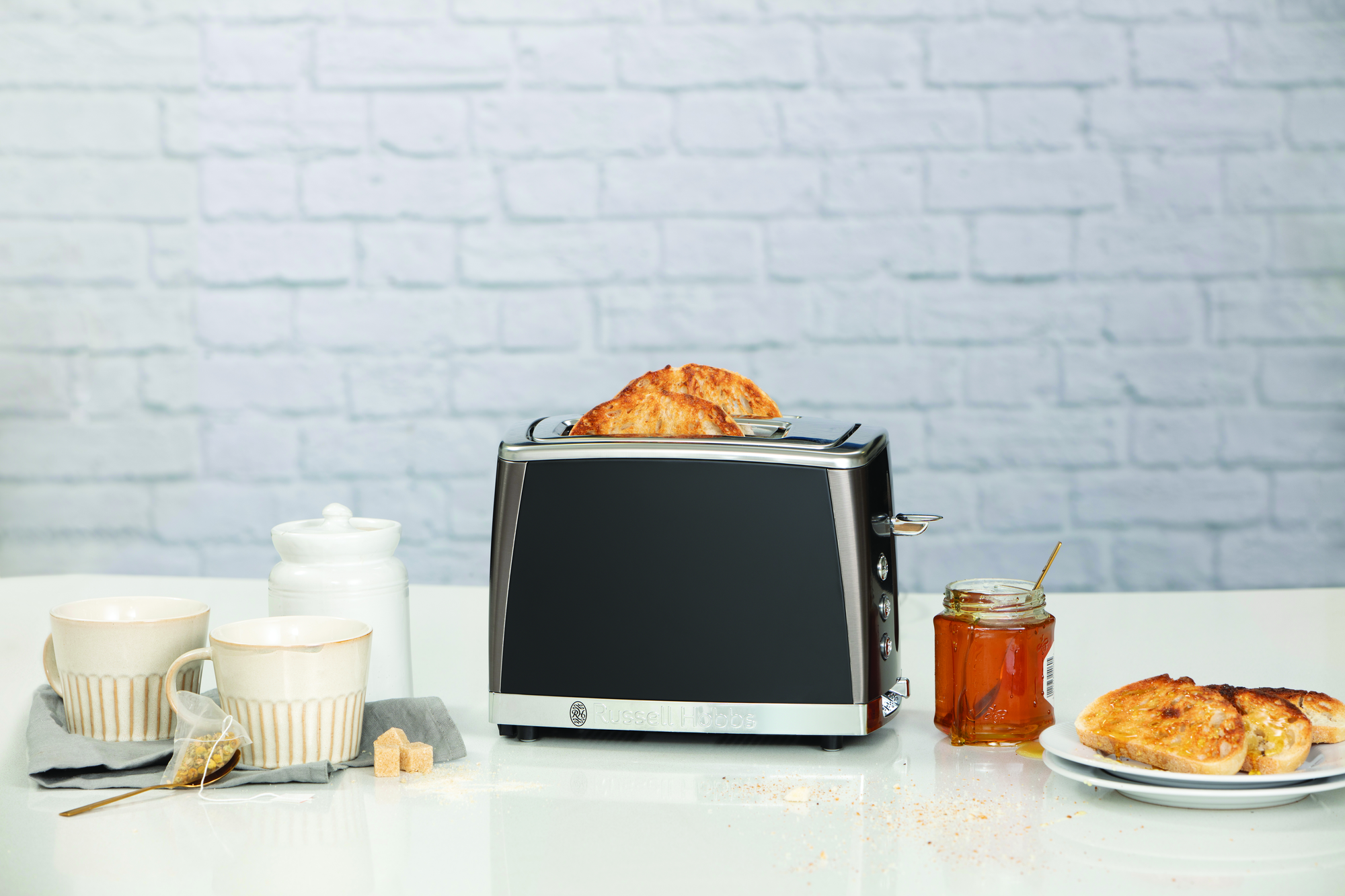 Toaster with croissants, a jar of honey, and tea cups on a white surface against a brick wall.
