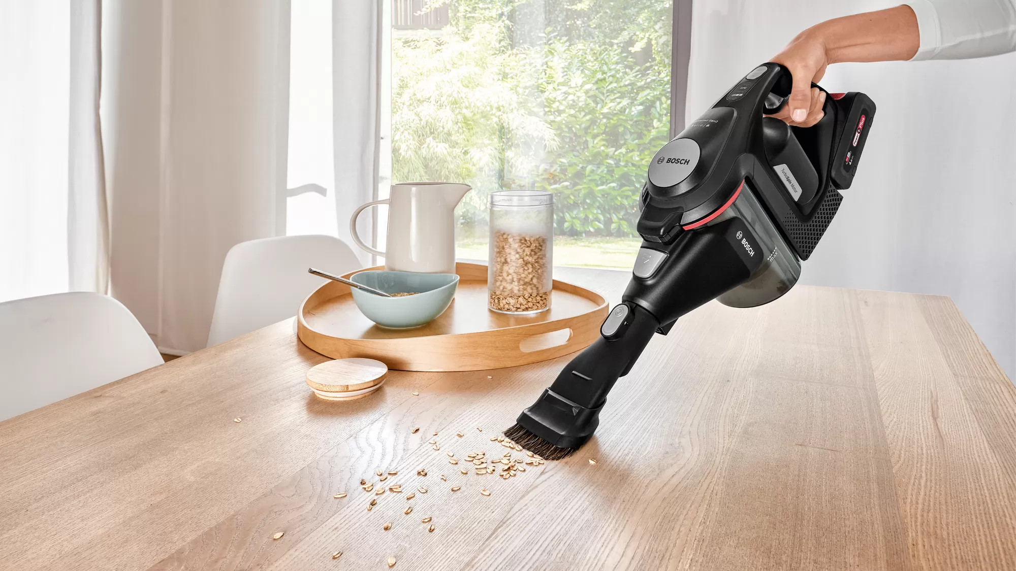Een zwarte en rode draadloze stofzuiger die een houten tafel schoonmaakt. Er zijn wat korrels op de tafel gemorst.