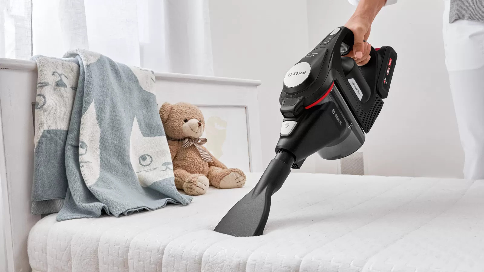Een persoon stofzuigt een wit bed met een zwart-rode Bosch stofzuiger, teddybeer en deken.