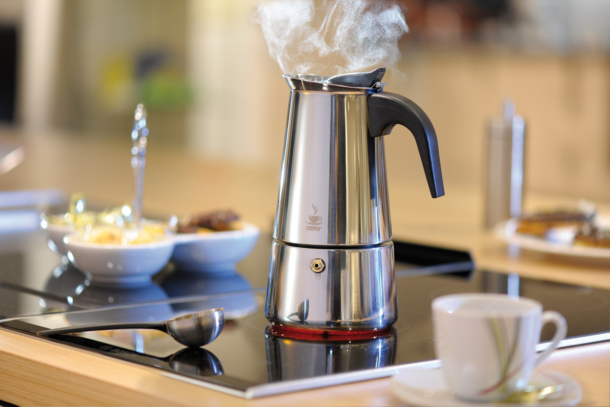 Ein silberner Espressokocher auf dem Herd mit aufsteigendem Dampf, einer Tasse und einem Löffel auf schwarzer Oberfläche.