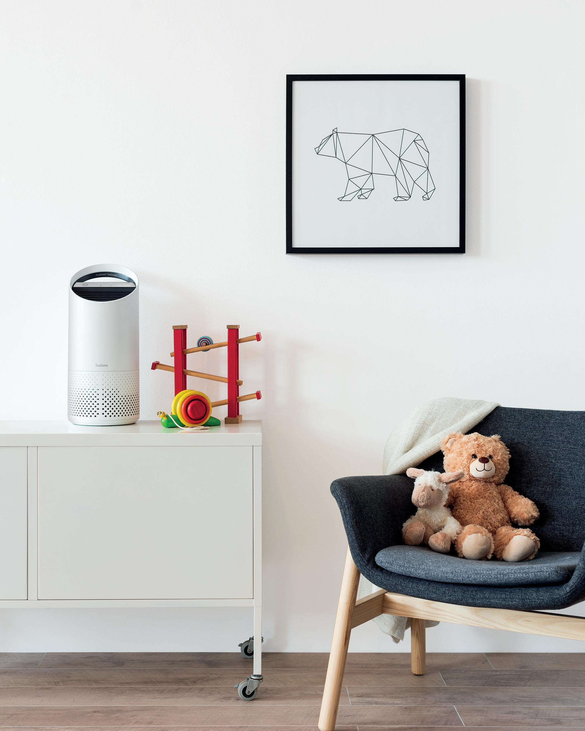 Ein Zimmer mit weißem Schrank, Luftreiniger, rotem Spielzeug, einem gerahmten Bärenbild und einem Teddybären auf einem Stuhl.